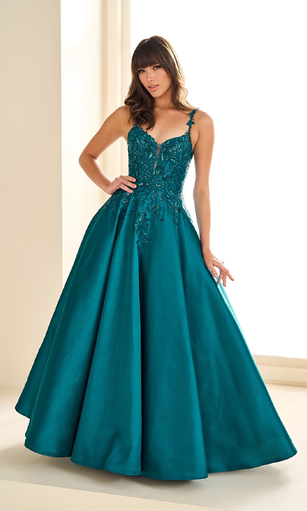 A woman stands indoors by a window wearing the Ellie Wilde EW36094, a long prom dress featuring a teal beaded corset bodice and full brushed satin skirt.